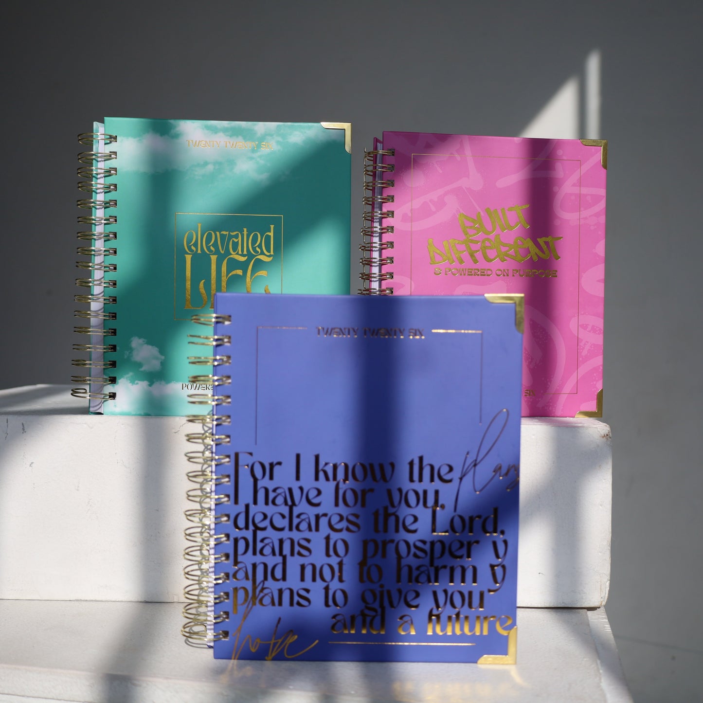 Three spiral-bound notebooks in blue, pink, and green on a white surface with a gray background.
