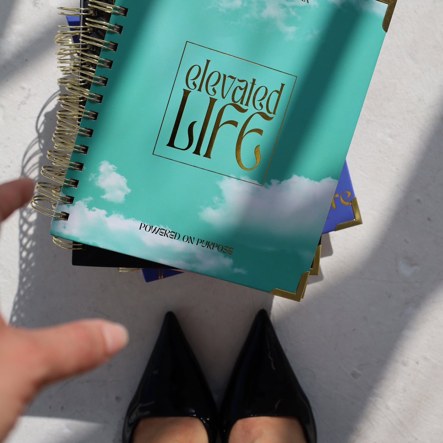 Teal spiral-bound book titled 'Elevated Life' on a white surface with a hand pointing at it.