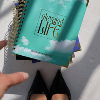 Teal spiral-bound book titled 'Elevated Life' on a white surface with a hand pointing at it.