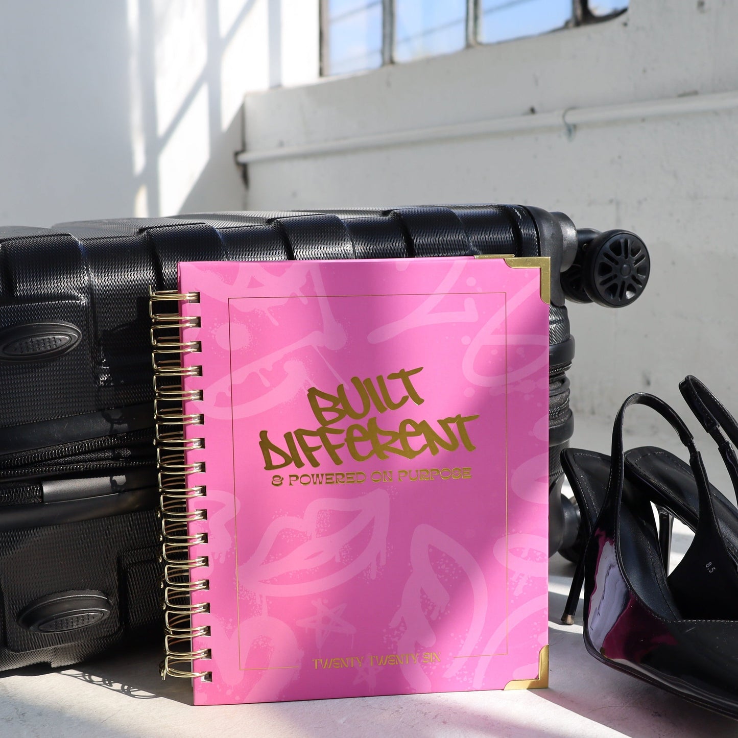 Black suitcase with a pink notebook titled 'Blunt Aftercut' and black high heels on a white floor.