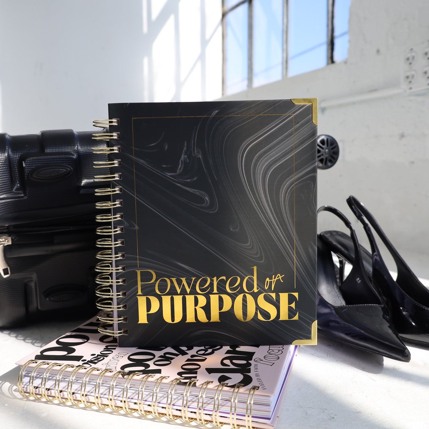Notebook titled 'Powered by Purpose' on a surface with shoes and a handbag in the background