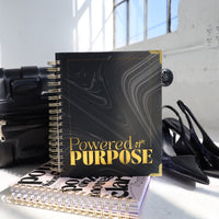 Notebook titled 'Powered by Purpose' on a surface with shoes and a handbag in the background